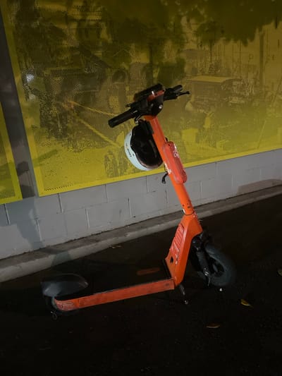 A bright orange electric scooter is parked on a dark asphalt surface, leaning against a light grey brick wall. The scooter has a black helmet attached to its handlebars, with a white outer shell and black interior. A red reflector is mounted on the stem of the scooter. Behind the scooter, a yellow perforated metal fence is visible, with an abstract pattern of darker shapes that appear to be reflections or shadows of nearby structures. The overall scene is dimly lit, suggesting it is nighttime or an indoor space with artificial lighting. The location is identified as Brisbane, Australia, although no specific landmarks are discernible in the image.