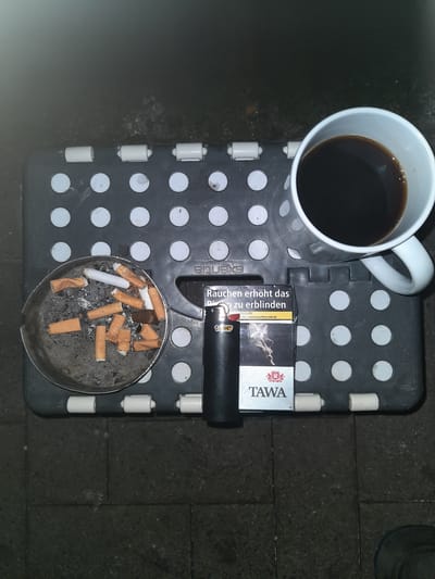 In the city of Celle, Germany, an overhead shot depicts a small, dark grey portable stool with a textured surface featuring a pattern of circular indentations. Resting on the stool are several objects. To the left, a round, metallic ashtray is filled with numerous cigarette butts, some with orange filters and others with white filters, along with ash and debris. Next to the ashtray, a black lighter lies horizontally. To the right of the lighter, a pack of "TAWA" cigarettes is positioned vertically, displaying a warning label in German that reads "Rauchen erhöht das zu erblinden" (Smoking increases the risk of blindness) and an image of smoke. To the right of the cigarette pack, a white ceramic mug is filled with dark liquid, presumably coffee. The stool is placed on a dark, textured surface, possibly pavement or an outdoor floor. The lighting suggests it is nighttime or a dimly lit environment.