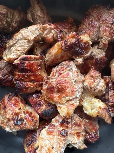 The image shows a close-up, overhead view of many pieces of cooked meat, likely steak or pork, piled in a dark pan. The meat is seared with grill marks on some pieces and has a rich, brown, and reddish-pink hue, indicating it is well-cooked. The lighting suggests an indoor setting, possibly a kitchen or a restaurant. There are no people or specific environmental details visible that would allow for the pinpointing of a location within Подолино Ст, Russia.