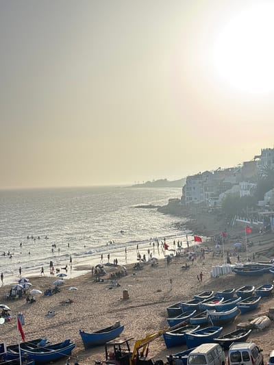 This image depicts a bustling beach scene in Taghazout, Morocco, at what appears to be late afternoon, judging by the low-angle sunlight casting long shadows. The ocean is active with numerous people swimming, surfing, and wading in the water. Along the shoreline, a sandy beach is populated with sunbathers, beachgoers, and a collection of blue fishing boats of various sizes, some resting on the sand and others closer to the water's edge. Several red flags are planted in the sand, some attached to poles, and beach umbrellas are scattered throughout the scene, providing shade.
In the background, the town of Taghazout is visible, characterized by white and light-colored buildings cascading down a hillside towards the coast. Some buildings have multiple stories and balconies. A rocky coastline is also present, meeting the ocean. The sky is hazy, possibly due to mist or the setting sun, with the sun itself visible as a bright, diffused light source in the upper right portion of the frame.
On the beach, there are various activities: people are walking along the shore, sitting under umbrellas, and relaxing on the sand. Some children are visible playing near the water. A small yellow excavator is parked on the sand, adjacent to some of the boats. White vans and other vehicles are also present on the beach area, near the boats. The overall atmosphere is one of a lively coastal resort town enjoying a day at the beach.