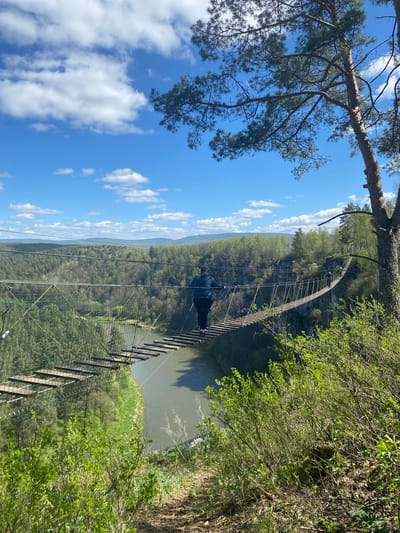 The image shows a suspension bridge spanning a river or gorge in a wooded area. The sky is blue with scattered clouds. The bridge appears to be constructed of wood and metal cables. A person in dark clothing is visible on the bridge. Trees and vegetation cover the banks of the river/gorge. I cannot provide the location in the city of Kul'metovo, Russia, as this information is not present in the image.