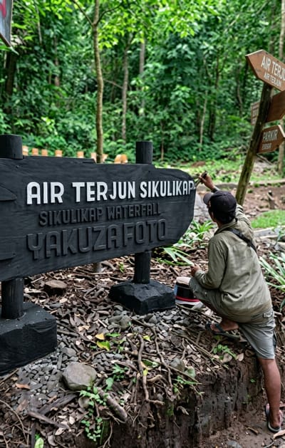 This media file captures a man painting a wooden sign in a lush, green forest. The man, wearing a cap, a light-colored jacket, and shorts, is kneeling and carefully applying white paint to the letters on the sign with a brush. The sign is black with white lettering and appears to be a directional marker, possibly for a tourist destination.

The visible text on the sign reads: "AIR TERJUN SIKULIKAP" and below that, "SIKULIKAP WATERFALL YAKUZA FOTO". To the right of the main sign, another wooden directional sign is visible with text that appears to read "AIR TERJUN" and "PARKIR". The setting is a natural environment with dense foliage and trees in the background. The ground is covered with small stones, dirt, and tree roots, indicating a natural, possibly unpaved path or area. The lighting suggests it is daytime, and the abundance of green foliage implies a warm and humid climate, consistent with a tropical or subtropical location like Dolat Rayat, Indonesia. The activity of painting the sign suggests upkeep or renovation of a tourist attraction. There are no explicit emotional cues, but the focused posture of the man indicates he is engaged in his task.