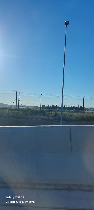 The image depicts an outdoor scene with a clear blue sky. A tall street light with a modern-looking fixture is visible on the left side of the frame. In the background, a chain-link fence runs along the horizon, separating the foreground from a grassy area with scattered vegetation and trees. The right side of the image shows a white surface with some streaks or stains. The location is possibly in or near Quart de Poblet, Spain. There are no people visible in the image. The photo was taken on a Galaxy M52 5G on 21 May 2026.