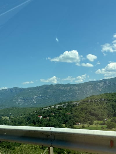 The image shows a landscape with mountains in the background and lush green trees in the foreground. There are a few small houses scattered among the trees, suggesting a rural setting. A metal guardrail runs along the bottom of the image, indicating that this view might be from a road or an elevated viewpoint. The sky is blue with a few scattered white clouds. The scene appears to be captured during the daytime under clear weather conditions.