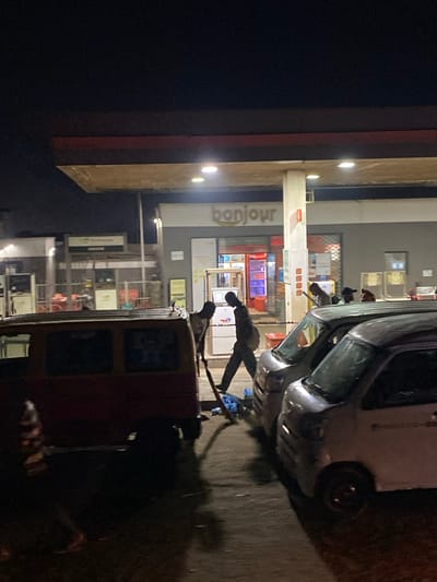 A brightly lit gas station at night. Above the pumps, the sign reads "bonjour". Several cars are parked in front of the gas station, and it appears that people are putting gas in their vehicles. The scene is captured at an angle, with the foreground being dark and a bit blurry.