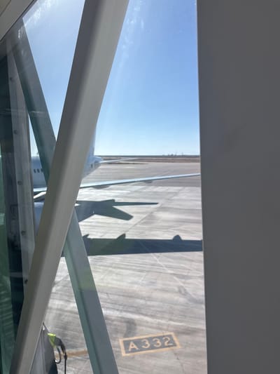 The image shows a view from an airport jet bridge looking out onto an airplane parked on the tarmac. The jet bridge structure, composed of white metal beams and glass panels, is partially visible in the foreground, creating a frame for the scene. The airplane, predominantly white with its wings extended, is positioned on a concrete surface marked with yellow lines. A shadow of the airplane's wing is cast on the tarmac. In the lower right corner of the image, a yellow painted number "A332" is visible on the tarmac. The sky is clear and blue, suggesting a sunny day. The scene is set at an airport in Mellita, Tunisia, indicated by the context of the image.