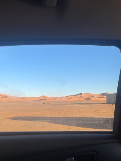 A view from inside a vehicle, showcasing a desert landscape through the open window. The sky is a clear blue. Adjacent to the open window is a ridge of sand dunes. A sandy expanse stretches to the horizon, interspersed with dark lines, likely shadows. The interior of the vehicle, including the frame of the window, is visible in the foreground. A Ford logo is seen on the glass.