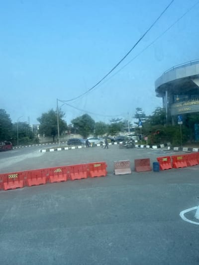 The image depicts a street scene in Serang, Indonesia, possibly near a transportation hub. In the foreground, a row of bright orange traffic barriers with black and yellow chevron markings stretches across the frame. Behind these barriers, the asphalt road surface is visible. Further back, several vehicles are present, including at least one car and what appears to be a three-wheeled vehicle. People are also seen walking in the area.
Dominating the background is a modern building with a curved facade, possibly a terminal or government building, as suggested by signage that partially reads "TERMINAL PAKUPATAN SERANG". The sky above is a clear, pale blue. Electrical wires are strung across the sky. Trees and greenery are interspersed around the area, indicating an urban environment with some landscaping. The overall impression is of an outdoor public space with ongoing activity.