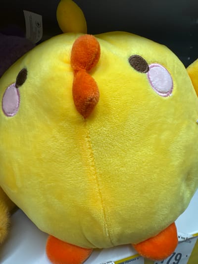 The image shows a close-up, overhead view of a yellow plush toy, possibly a duck or chick, lying on a white surface. The toy is predominantly yellow with orange accents on its beak and ears. It has embroidered pink cheeks and dark brown eyes. The plush material appears soft and fuzzy. There are labels visible on the toy, including one with a small airplane icon and some unreadable text, and another on the right side of the image with a black number "79" and some Cyrillic text. The lighting suggests an indoor setting. The image is taken from a high angle, looking directly down at the toy. There are no people or discernible activity taking place, it is a static shot of the toy.