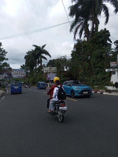 The scene depicts a multi-lane paved road in Taohom, Laos, under an overcast sky. In the foreground, two individuals ride a dark-colored motorcycle (license plate S 5813 XO) away from the viewer. The driver wears a yellow helmet and a red shirt. The rear passenger wears a white t-shirt with a black star graphic, grey pants, and a black backpack, and is barefoot. In the left lane, ahead of the motorcycle, a blue minivan is visible. In the opposing right lane, a bright turquoise SUV with a black roof (license plate D 1032 TAD) approaches. The road is lined with dense green foliage, including numerous palm trees, and various buildings. Several commercial signs are visible in the background, including a large sign for "SINDANGLAYA HOTEL & RESTAURANT" and a smaller "Canon digital shop" sign on the right. Overhead power lines span the road. Additional vehicles, primarily motorcycles and another blue minivan, are visible further down the road.