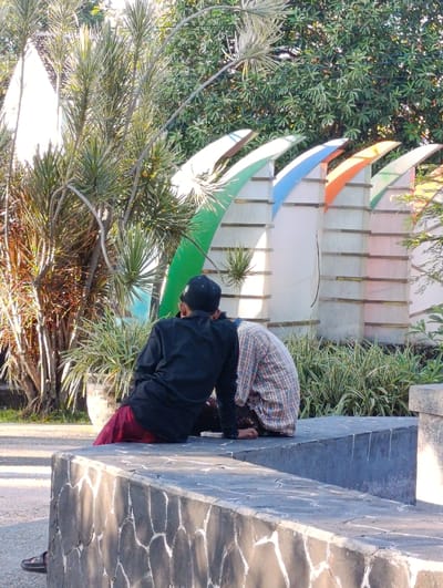 Two men are sitting on a concrete bench in a park in Kecamatan Asembagus, Indonesia, located at latitude: -7.75, longitude: 114.218. The men are facing each other, with their legs crossed. The man on the left is wearing a black shirt and a black cap. The man on the right is wearing a plaid shirt. They are both looking at something off-camera. Behind them is a row of colorful, decorative panels with curved tops, made of white material. To the left of the bench are palm trees and a concrete planter. To the right of the bench is a concrete structure. The background is a lush green landscape, with trees and bushes.