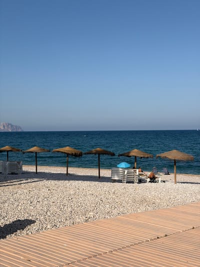This media file depicts a serene beach scene on a clear, sunny day, likely in the late afternoon given the long shadows. The setting is a beach lined with light-colored pebbles, with a wooden walkway running along its edge. Several thatched umbrellas are erected, providing shade for rows of empty white lounge chairs. In the background, the calm, deep blue sea stretches to the horizon, with a faint outline of a mountainous coastline visible in the distance. The sky is a clear, pale blue, suggesting fair weather.

There are a few people present, seemingly relaxing on the beach. One individual is visible sitting on a lounge chair under an umbrella, and another person is lying on a chair further back. A small, bright blue umbrella stands out against the natural tones of the scene. The overall atmosphere is one of tranquility and relaxation, characteristic of a beach resort. No visible text is present in the image. The orientation of the image is rotated 90 degrees clockwise.