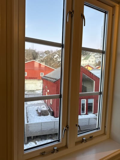 The image displays an outdoor scene in Stamsund, Norway, viewed through a white-framed window featuring two vertical panes, two horizontal muntins, silver latches, and lower stays. A retractable insect screen is partially visible within the upper window frame.
The environment is a winter landscape, characterized by snow covering the ground, rooftops, and rocky hills in the background. The sky is clear and blue.
In the midground, multiple buildings are visible. To the left, a large red building with a white garage door and two small white square windows is situated. Adjacent to it, a smaller red wooden building with a dark-pitched roof and a white window is present. Further to the right, another red wooden building with a steep, angled roof and lower windows, including a door, stands. Beyond these, uphill, a multi-story white building with a green roof and a yellow building are partially visible.
The outdoor area directly below the window features a snow-covered deck or platform, bordered by a white slatted railing. On this deck, a picnic table and benches are buried under snow. Rocky, snow-dusted hills with bare trees rise in the background, and a tall communication mast or tower is visible on the right horizon.
No individuals, animals, or active interactions are present within the scene. The lighting suggests daytime conditions.