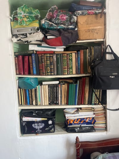 This is a photograph of an interior scene depicting a makeshift shelving unit filled with various items, predominantly books. The shelving unit is integrated into a white-painted wall, suggesting it might be a built-in feature or a custom addition. The overall impression is one of cluttered organization.
The top shelf contains several fabric bags with floral patterns, a brown cardboard box, and a dark blue duffel bag. Beneath this, a layer of books is visible, some with red covers.
The second shelf from the top holds a collection of books with predominantly dark spines, some appearing to be in Arabic script. A black shoulder bag with a silver zipper is prominently placed on the right side of this shelf.
The third shelf is filled with more books, varying in color and size. A bright teal-colored bag is positioned on the left side of this shelf.
The fourth shelf also contains a dense arrangement of books, with some spines featuring Arabic text.
The bottom visible shelf has a black storage box with a clear glass bowl on display. Above this box, other items are stacked, including books and what appears to be a blue plastic bag with orange lettering.
The environment appears to be a room with white walls. The lighting suggests it could be daytime, possibly with artificial lighting contributing to the overall illumination. The presence of books indicates a potential study or living area. The provided context mentions the location as Tashlak, Kyrgyzstan, suggesting this is a domestic interior within that city. The photograph captures a static scene, with no discernible actions or interactions taking place.