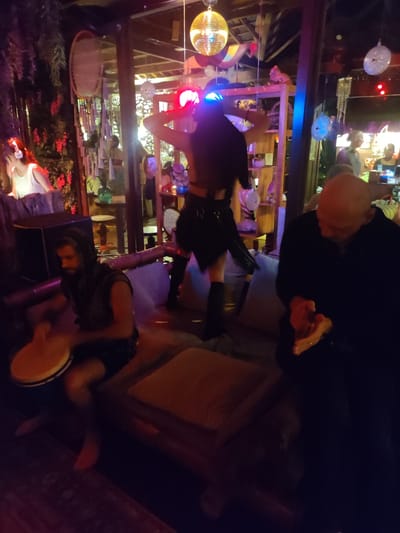 The image depicts an indoor nightlife scene, possibly a bar or lounge, in Lokaserena, Indonesia. Dim lighting is evident. A DJ with illuminated headwear stands on a raised platform in the center of the frame, apparently dancing with hands raised. A man plays a drum, seated on a cushioned surface to the left. To the right, a bald man is seated, looking down and rubbing his hands together. Other patrons can be seen in the background, engaging in conversation. Decorative elements include hanging lights, greenery, and other adornments. The overall atmosphere appears to be relaxed and celebratory.