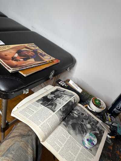 The image captures a detailed, high-angle view of a cluttered indoor space, likely a corner next to a wall or a bedside area. The scene is static, showing various personal items arranged on different surfaces.

On the left side of the frame, a black, hard-shelled case, possibly for a musical instrument or a briefcase, rests against a plain white wall. It features silver-colored latches. Stacked on top of this case are several magazines. The top magazine is clearly visible, titled "PHOTO MAGAZINE" (the word "MAGAZINE" is partially obscured, but "PHOTO" is clear). Its cover features a close-up portrait of a shirtless man with a blue headwrap and a prominent, multi-strand necklace.

To the right, a surface, possibly a table or shelf, is densely packed with items. A large book or magazine is open, displaying two pages with French text and black-and-white photographs. The left page features an image of an old utility vehicle, possibly an ambulance, surrounded by what appear to be barrels or large cylindrical objects. The right page shows a crowd of people, some holding buckets, in what looks like a historical or documentary photograph. Part of the text "le so" is visible at the bottom of the left page, possibly from a newspaper or periodical title.

Near the open book, a small, white ceramic dish serves as an ashtray, containing several cigarette butts and a small green object, possibly a plant or piece of foliage. Next to this dish lies a black remote control with numerous buttons. Behind the remote, a tiny potted plant in a white pot is partially visible. Further back on the table, several smoking-related items are present: a black and silver box with "8 ORIGINAL" visible, and another cigarette pack with the warning "Fumer diminue" (Smoking reduces/decreases). A small white box, possibly a matchbox or another cigarette accessory, has the text "DEPUIS DES ANNÉES" (for years/since years) printed on it. A small, colorful, spherical or circular decorative object with blue, green, and pink patterns rests on the open book on the right side.

The background on the upper left is a plain white wall. The lower right portion of the image shows a wooden floor and a grey, textured fabric, possibly part of a cushion or clothing, along with a silver-colored metal leg or stand of furniture. The overall impression is one of a lived-in space, suggesting the interests and habits of its occupant, such as reading and smoking. The visible French text suggests a Francophone context for the items and possibly the location.