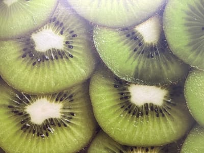This is a close-up, overhead shot of several sliced kiwis. The slices are arranged in a way that their circular, vibrant green flesh and characteristic black seeds are visible. The skin of the kiwis, with its fuzzy texture, is also partially shown. The image appears to be taken indoors, possibly on a flat surface. There are no people or animals present, and no indication of a specific location in Moscow, Russia. The lighting is somewhat diffused, and there are minor imperfections or scratches visible on the surface, suggesting it might be a printed image or a decorative surface.