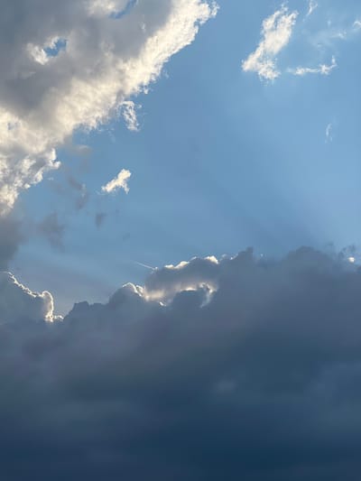 The image shows a sky with clouds and sunbeams. The location is the city of Straubing, Germany. In the upper left quadrant, there are large, white clouds with bright edges, indicating sunlight hitting them. The rest of the sky is a clear blue, with lighter patches suggesting sunlight filtering through other clouds. In the lower half of the image, there are darker, more voluminous clouds that obscure much of the sky. Sunbeams are visible emanating from behind these clouds, creating dramatic light rays across the blue sky. Near the center of the image, a faint white contrail from an aircraft is visible. No people, subjects, or specific objects are discernible within the scene, other than the natural elements of the sky. The scene appears to be an outdoor environment, likely at a high vantage point given the view of the sky.