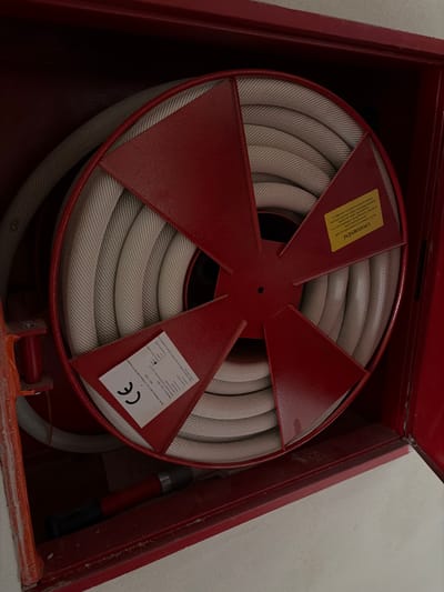 This is a close-up, eye-level photograph of a coiled fire hose inside a red metal fire hose cabinet. The cabinet door is open, revealing the hose reel, which is red with five spokes. The hose is light gray and tightly wound around the reel. A white sticker with a black "CE" mark is visible on the reel. To the right of the "CE" sticker, there is a yellow sticker with text in an unknown language. The lower portion of the cabinet contains a nozzle for the fire hose, which is red and black. The fire hose cabinet is mounted on a light-colored wall, likely concrete or plaster. The image appears to have been taken indoors, possibly in a public building or hallway. The specific location in Příbram, Czechia is not discernible from the image itself.