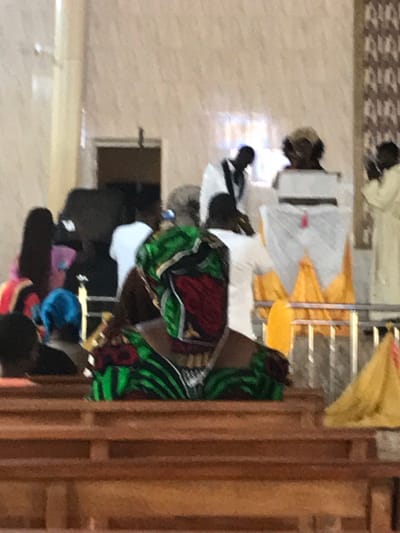 The image captures an indoor gathering in Adum Afo, Nigeria. In the foreground, a dark-skinned individual, likely a woman, is seated facing away from the camera, wearing a vibrant green, black, and red patterned headwrap and matching garment. Wooden pews are visible in the foreground and mid-ground. Further back, multiple dark-skinned individuals are present. One man in a white robe with black trim is seen in the mid-background, facing towards a central area. Another person with light-colored hair or a head covering is partially visible behind a white-draped structure, possibly a podium, adorned with a red detail and situated above yellow fabric. To the far right, a person in a light-colored robe is partially visible. Metal railings separate sections of the room. The background features light-colored, possibly tiled walls, with natural light filtering through windows. A vertical patterned wall strip is present on the right.