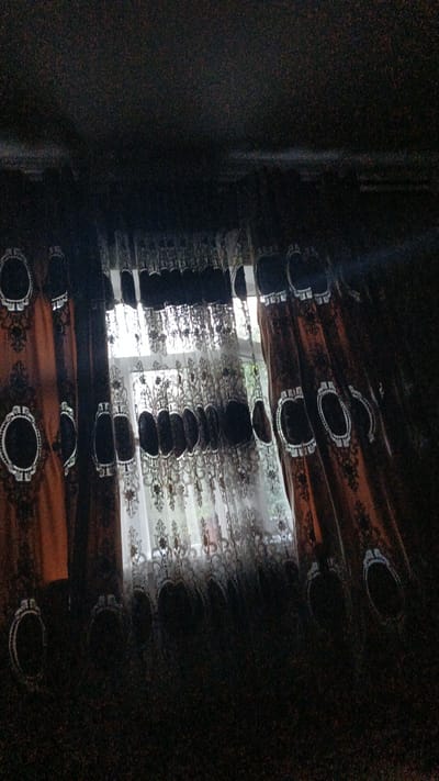 The image shows a view through a window with ornate curtains partially drawn. The curtains are made of two layers: a darker, patterned fabric on the outside and a sheer, lace-like fabric underneath. The outer curtains are a dark brown or rust color with a black lace overlay featuring an oval motif. The inner curtains are white or off-white lace with floral patterns. Sunlight filters through the lace, creating a diffused illumination. Outside the window, greenery and a hint of what appears to be a building can be seen, suggesting an outdoor environment. The overall lighting is dim, with the window area being the brightest. The image is taken from inside a room looking outwards. The location is identified as Navobod, Tajikistan.