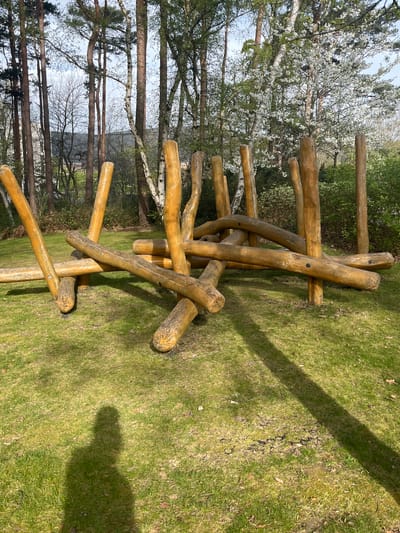 The image shows a wooden structure on a grassy area in the Borough of Runnymede, United Kingdom. The structure appears to be a play feature, constructed from unpolished logs arranged in a haphazard, climbing-frame-like configuration. The logs are various lengths and are attached to each other with black screws, forming a series of angled supports and horizontal bars. The surrounding environment consists of a mowed grassy area with trees bordering the edge of the image. A few of the trees are in full bloom, displaying white flowers. Shadows from the structure and trees are cast onto the grass, indicating the presence of sunlight. The sky is partly cloudy. There are no people or actions directly visible in the image.