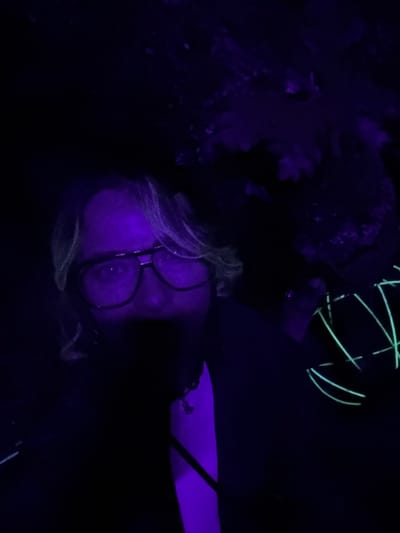 A single individual is centered in a darkly lit environment within the city of Bukovik, Montenegro, illuminated primarily by ultraviolet light. The subject, an adult with light-colored hair and dark-framed glasses, gazes directly forward. Their face and skin exhibit a purple luminescence, with the lower facial features obscured by shadow. A subtle necklace is visible on their chest. The individual holds an object in their right hand (viewer's left), which consists of neon-green glowing wires arranged into a stylized, mask-like pattern. The background is indistinct and deeply shadowed, revealing faint purple-hued textures on irregular surfaces, suggestive of rough terrain or natural formations.