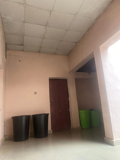 The image depicts an interior scene, likely a room or hallway in Wauro Leggal, Nigeria. The walls are painted in neutral tones, with a section of tiled wall on the left. Above, a bright light source suggests daytime or a well-lit interior space.

Prominent objects in the scene include two large, black cylindrical bins positioned vertically on the floor. In an elevated alcove, two bright green plastic containers are stacked. A dark wooden door or panel is visible on the wall, partially obscured by the alcove. There is a small, square electrical outlet on the peach-colored wall.

The scene appears to be a storage area or a utility space within a building. There are no people visible, and no specific activity is taking place. The setting is utilitarian, with an emphasis on practical storage solutions. The overall impression is one of a simple, functional interior. No text is visible in the image.