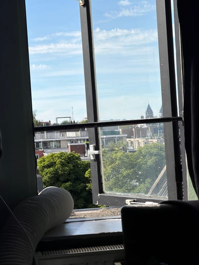 The image captures a view from a window in Amsterdam, Netherlands. The window is open, revealing a cityscape dominated by apartment buildings and lush green trees. The sky above is a clear blue with scattered white clouds. In the foreground, a white, flexible tube is visible, likely an air conditioner hose, resting on a window ledge. A radiator is also partially visible beneath the ledge. The architecture of the buildings suggests an urban residential area. There are no people visible in the frame.