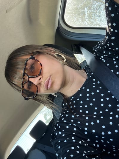 The image shows a woman with brown hair and bangs wearing large, brown-rimmed glasses and a black and white polka dot dress. She is in the passenger seat of a car, indicated by the seatbelt across her body and the car's interior visible in the background, including part of the sunroof and the back of a headrest. The woman is looking at the camera with her lips puckered. The lighting suggests it is daytime, with sunlight streaming in from the car's sunroof. No specific location in Le Muy, France, can be identified from the image.