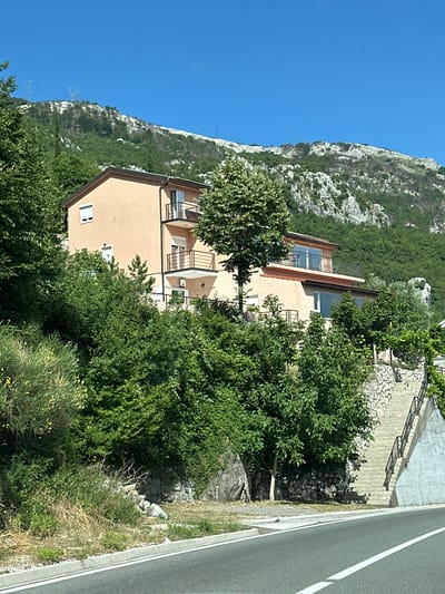 The image depicts a multi-story house situated on a hillside in Vrela, Montenegro. The house is painted a light orange and features multiple balconies with dark railings. It is surrounded by lush green vegetation, including trees and shrubs. In the foreground, a paved road with white lane markings curves to the right. To the right of the house, a stone-walled staircase ascends the hillside. The background shows a steep, rocky mountain covered in dense green trees, with the clear blue sky above. Power lines and pylons are visible on the distant mountain slope. No people are present in the image.