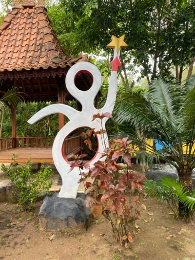 The image shows a white, abstract sculpture of a person reaching up with one hand, holding a gold star, located in Kecamatan Manyaran, Indonesia, at coordinates -7.826 latitude and 110.816 longitude. The sculpture is standing on a large, black rock in a patch of dirt. There is a small, wooden gazebo to the left of the sculpture. Lush green foliage surrounds the gazebo, and a red-leafed bush grows in front of the sculpture. A palm tree to the right of the sculpture has fronds reaching into the frame. The sculpture has red trim around the holes cut out for the arms, legs, and head. The sculpture is in a bright outdoor setting with a bright blue sky in the background.