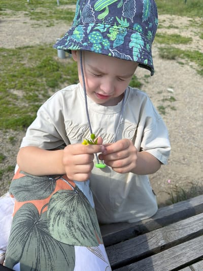 A young boy wearing a patterned bucket hat and a beige t-shirt is sitting on a wooden bench outdoors. He is holding a small yellow flower with green leaves in his hands, examining it with a focused expression. The boy's t-shirt has a subtle embroidered design. His left leg is covered by a pillow or cushion with a large green and orange leaf pattern. The background consists of a gravelly area with patches of grass. This scene is located in the city of Kul'dur, Russia.