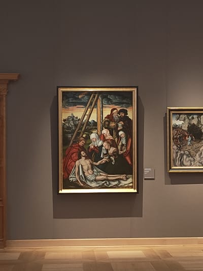 The image depicts an interior view of an art museum in Aschaffenburg, Germany. The scene is dominated by a large, vertically oriented framed painting centered on a dark gray wall. This painting, presented in an ornate gold frame, portrays a multi-figure religious scene, likely a Lamentation or Deposition of Christ. A pale, deceased male figure, partially clad in white drapery and exhibiting wounds on hands and feet, lies horizontally in the foreground. He is surrounded by at least six other figures, primarily women in various states of grief and support, dressed in period robes of red, white, green, and dark colors. One female figure cradles the head or upper torso of the deceased, while another kneels near his feet. In the background, a wooden ladder leans against an upright beam, under a dramatic, cloudy sky. Distant hills and a structure resembling a fortified building or castle are visible on the left.
To the right of the main painting, a smaller, horizontally oriented framed painting is partially visible, also in a gold frame. This artwork depicts a dense crowd of small figures within a rugged, hilly landscape featuring trees and rocky formations, under a similarly overcast sky.
The museum wall is a uniform dark gray. A light wooden baseboard runs along the bottom of the wall, meeting a patterned wooden or tiled floor composed of light-toned rectangular and square segments. On the far left, a vertical wooden architectural element, possibly part of a doorway or furniture, is partially visible. No human visitors are present in the immediate museum space captured in the image.