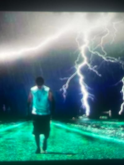 This image, set in the city of Zelenyi Hai, Ukraine, presents a dramatic, stylized outdoor scene. A single individual is prominently featured from behind, positioned centrally on a vividly green, grassy path or road that recedes into the distance. The person wears a light-colored sleeveless top, likely teal or light blue, and dark shorts, with their head obscured by shadow or distance. Above, the sky is dark, transitioning from deep blue to black, and is intensely illuminated by numerous bright white lightning bolts. These lightning strikes are thick, branching, and highly luminous, dominating the upper half of the composition and casting an ethereal glow over the environment. The overall impression is one of a powerful, perhaps ominous, natural event occurring within a highly saturated and visually striking landscape.