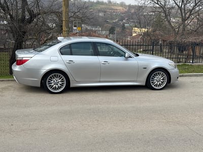 The image displays a silver four-door BMW E60 5 Series sedan parked on a light grey asphalt road in Profesor Ishirkovo, Bulgaria. The vehicle is oriented with its left side towards the viewer, facing right, and appears to be in good external condition, equipped with multi-spoke alloy wheels and a subtle rear lip spoiler. The road surface exhibits minor cracking. To the immediate right of the vehicle, a concrete curb leads to a patch of sparse grass and a black metal fence. Behind the car, a utility pole with an attached electrical meter box displaying "1152" is visible, situated among a dense cluster of bare deciduous trees. Further in the background, residential buildings with tiled roofs are discernible, with vegetated hills rising in the distance under an overcast sky. No individuals, actions, or interactions are present in the scene.