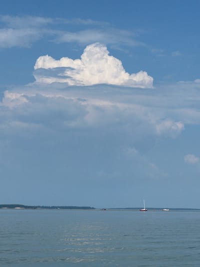 The image depicts a vast body of water under a bright blue sky filled with dramatic, fluffy white clouds. In the distance, a thin strip of land with dense, dark green vegetation stretches across the horizon. A few small boats are visible on the water. One is a red and white sailboat with its mast prominently displayed. Another appears to be a white motorboat. The water is a calm, muted blue-gray, with subtle ripples reflecting the light. The overall impression is one of a serene, open landscape. The image is undated and location is not specified.
