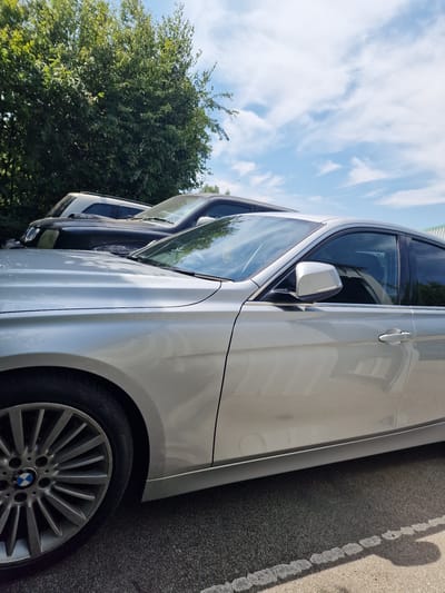 In Fenland District, United Kingdom, on a bright, sunny day with scattered clouds, a silver BMW sedan is parked on an asphalt surface. The car is seen from the side, with its front passenger door and fender in focus. The vehicle has a distinctive alloy wheel with the BMW logo prominently displayed. To the left of the BMW, partially visible, is a black SUV. Behind the cars, a dense line of lush green trees forms a natural backdrop, with the sky visible above. The scene appears to be an outdoor parking area.