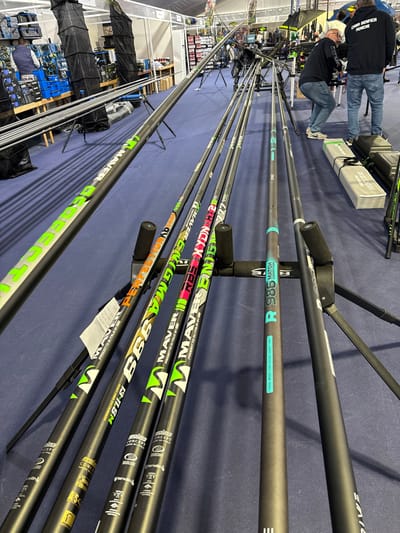 The image depicts an indoor fishing tackle exhibition booth located in Mora, Portugal. The foreground is dominated by a display of ten carbon fiber fishing poles, horizontally aligned on black stands with foam cradles. The poles are predominantly black with various brand names and model numbers, including "MAVER", "BBB", "PERFECTION", "XYDON HT2", "RBS", and "R985 MATCH COMPETITION", rendered in vibrant green, orange, pink, and light blue lettering.

The floor is covered with dark blue carpeting. In the mid-ground, two adult males are visible. One man on the left wears a blue vest over a dark shirt. The second man, to the right, wears a black hoodie with "CASA BENFICA PESCA" in white text on the back, and blue jeans; he is bent over, examining items on a low display.

The background shows a temporary exhibition space with white wall panels and a white tent-like ceiling structure. Additional fishing equipment is displayed, including stacks of blue storage boxes, several black fabric rod bags, shelves stocked with smaller items, and green and black fishing chairs, one bearing the label "BEASTMASTER". More fishing poles and tripod stands are visible further back.