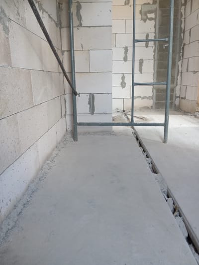 The image depicts an interior view of a building under construction. The walls are constructed from light-colored concrete blocks, some of which have visible mortar. Scaffolding, made of gray metal pipes, is erected in the center of the frame, supporting construction work. The floor is covered with a smooth, light gray concrete surface, and a visible groove or channel runs across it, containing debris. The lighting suggests an indoor environment with artificial light sources, creating shadows on the floor and walls. The scene appears to be a construction site in progress, with no people or specific identifiable objects beyond the building materials and scaffolding. No information is provided about the specific location in Terema, Russia, within the image itself.