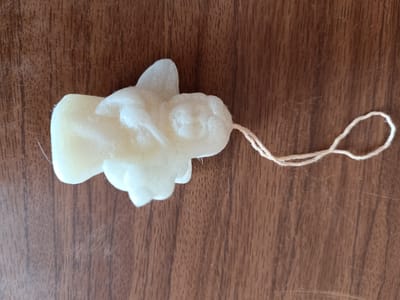 A pale yellow, angel-shaped object, possibly a soap or candle, rests on a brown wooden surface. The object has a string loop attached to its right side. The wooden surface has a visible grain pattern with vertical lines. The lighting suggests an indoor environment. There are no people present. The image does not provide any information about the city of Tebras, Latvia.