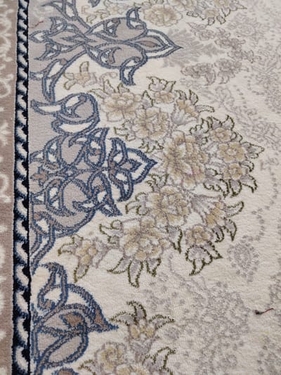 The image shows a close-up view of an intricately patterned rug. The rug has a cream-colored base with a repeating floral and vine motif in shades of beige, grey, and muted blue. The border of the rug, visible on the left side of the image, features a black and white zigzag pattern, with a beige strip adjacent to it. The rug appears to be made of a soft material, with individual fibers visible upon close inspection. There is a small, red thread visible on the right side of the rug, appearing to be a loose fiber. The lighting suggests an indoor environment, with no specific details of the surroundings or location provided within the image itself. Therefore, it is not possible to determine its location within the city of Pitnak, Uzbekistan based solely on this image.