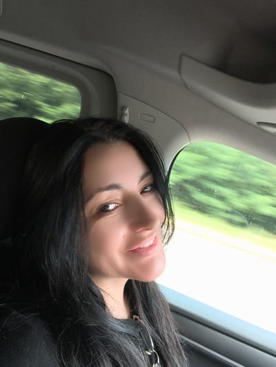 The image shows a woman in a car, looking at the camera and smiling. She has long, dark hair and is wearing a black top. The car interior is visible, including a gray headliner and a window. Outside the window, blurred green trees and a road are visible, suggesting the car is in motion. The lighting appears to be natural daylight. The scene is a close-up portrait of the woman within the context of travel.