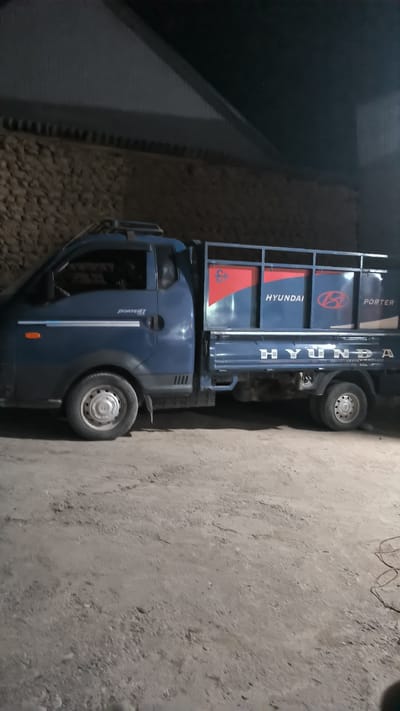 A blue Hyundai Porter truck is parked on a dirt or gravel lot at night. The truck has a silver rack on the back and the word "HYUNDAI" is visible on the side of the bed. The background shows a textured wall and a dark, sloped roof. The scene appears to be in a rural or industrial area, possibly on the outskirts of the city of Интернационал, Kyrgyzstan. There are no people visible in the image.