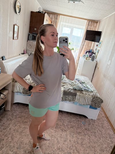 A woman is taking a mirror selfie in what appears to be a bedroom in a city apartment. She is wearing a gray t-shirt and light green shorts. She has a ponytail and is looking at her reflection. The room is cluttered with furniture, including a bed, a dresser, and a desk. There is a window with curtains in the background, and a television mounted on the wall. The floor is covered with a shag rug. Based on the provided context that this image is from Kul'dur, Russia, it can be inferred that this is a bedroom in an apartment in that city.