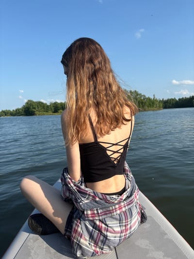 A young woman with long, wavy brown hair is sitting on a paddleboard on a calm body of water, facing away from the camera. She is wearing a black criss-cross back tank top, a plaid shirt loosely draped around her shoulders, and dark shorts. Her legs are crossed, and she is looking down and to her left. The background shows a shoreline lined with green trees and a clear blue sky with a few wispy clouds. The water is rippling gently, reflecting the sky and surrounding foliage. The location is not specified in the image but is assumed to be in Razdolnoye, Russia.