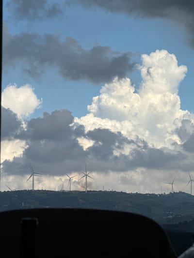 The image shows a view of a hilly landscape with wind turbines, under a sky filled with dramatic clouds. In the foreground, a dark, blurred object, possibly the interior of a vehicle, obscures the bottom portion of the scene. The middle ground features a green, undulating hill covered with vegetation and dotted with buildings. Several tall wind turbines are visible on the crest of the hill, their blades extended. In the background, the sky is a mix of bright blue and large, white, puffy clouds, interspersed with darker, more ominous clouds. There is a faint haze or smoke visible near the horizon, in the vicinity of some of the wind turbines, suggesting potential industrial activity or a fire. The image captures a sprawling, open environment, likely in a rural or semi-rural area with renewable energy infrastructure. While the prompt specifies the city of Đa Lộc, Vietnam, the visual information in the image itself does not contain specific landmarks or details that would confirm this location. The scene is naturalistic and atmospheric, with a focus on the interplay of light, clouds, and the man-made structures of the wind farm.