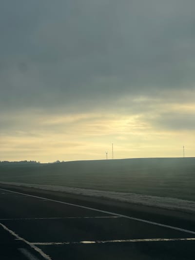 The image depicts an outdoor scene, likely in the Вольнаўскі сельскі Савет, Belarus area, given the context. The foreground shows a section of a road or paved surface with visible lines. Adjacent to the road is a field, possibly farmland, which extends towards the horizon. In the distance, several wind turbines are visible against a cloudy or hazy sky. The overall lighting suggests either sunrise or sunset.