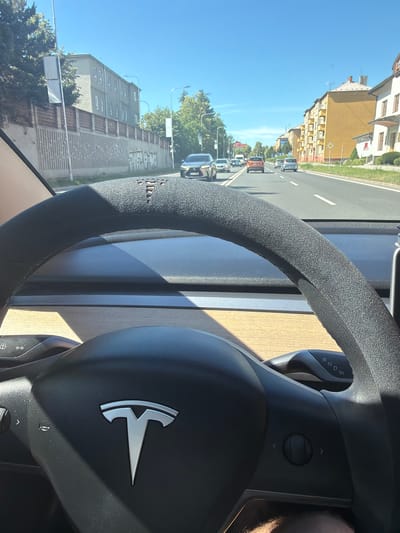 The image is a first-person perspective from inside a Tesla, looking out at a street scene and the car's steering wheel. The steering wheel is black with a textured grip and the Tesla logo in the center. A gear selector stalk, labeled "R N D P", is visible to the right of the steering wheel.

The scene outside the car shows a street with buildings and trees. The buildings appear to be residential and are mostly in shades of yellow and gray. There are other cars on the road, suggesting traffic. The sky is clear and blue, indicating a sunny day. A portion of the steering wheel shows wear and tear, with some of the material peeling off. The overall impression is a snapshot of a daily drive.

There is no visible text in the image that provides specific information about the location or time, other than the "R N D P" on the gear selector. The image was taken in Olomouc, Czechia. The lighting suggests it is daytime, likely late morning or afternoon due to the angle of the sun and the shadows cast. The atmosphere appears calm, with no indication of any particular event or strong emotions.