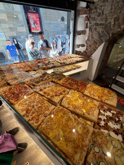 The image depicts a display of pizzas inside a bakery or shop. The pizzas are arranged on shelves and in a refrigerated display case. Various toppings are visible, including cheese, vegetables, and meats like sausage. In the background, through a glass window, several people are seen browsing outside the shop. Some are looking at a poster on the wall. The exterior wall of the building appears to be made of stone. The location is identified as Bergamo, Italy.